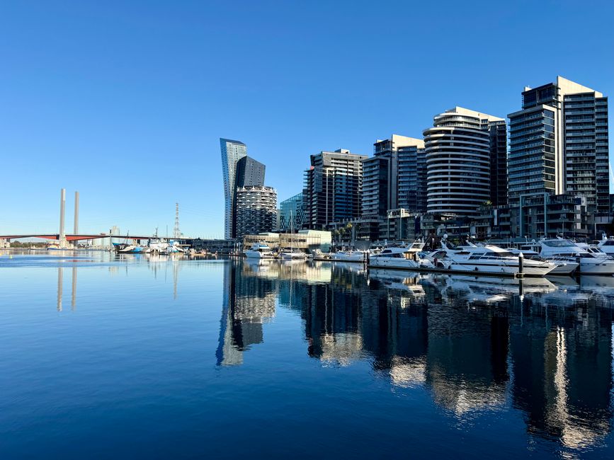 A panoramic view of Docklands featuring modern high-rise office buildings and residential flats made of glass and steel, reflecting sunlight and appearing clean and well-maintained. In the foreground, a marina with still water holding several white yachts and boats, their reflections clearly visible on the surface. The sky is bright blue with no clouds, and the scene is illuminated by natural daylight, emphasizing the sleek and tidy appearance of the waterfront area. The overall setting showcases a contemporary urban landscape with a focus on cleanliness and architectural detail, indicative of the professional cleaning services provided by Carpet Cleaning Docklands and aligned with the topic of avoiding common carpet cleaning mistakes in Docklands flats.