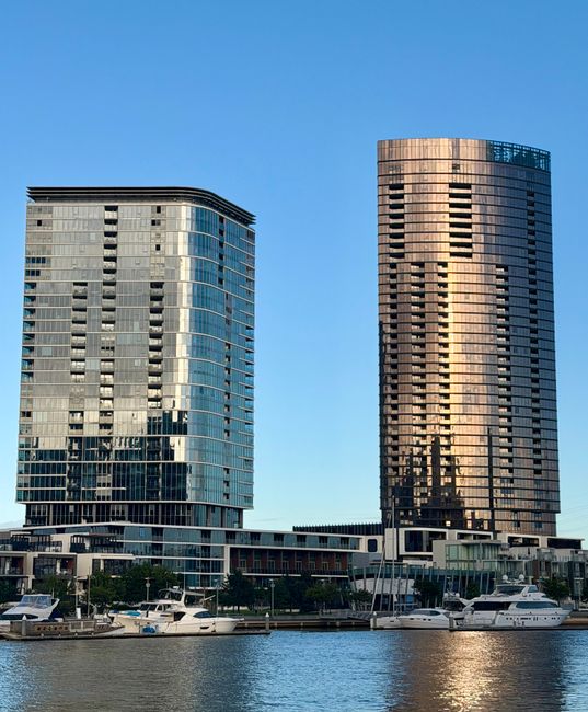 A daytime view of two modern high-rise office buildings situated along a waterfront in Docklands, with the left building featuring curved glass panels and reflective windows, and the right building displaying a grid-like pattern of dark and light windows. The buildings are predominantly constructed from glass and steel, reflecting the clear blue sky and surrounding structures. At the base, a row of yachts and boats are moored along the water's edge, with a promenade visible beneath the buildings. Bright natural lighting emphasizes the cleanliness and sleek appearance of the architecture. The scene underscores the importance of surface cleaning and maintenance for urban commercial properties, as highlighted by Carpet Cleaning Docklands, ensuring hygienic and visually appealing environments in the Docklands area.