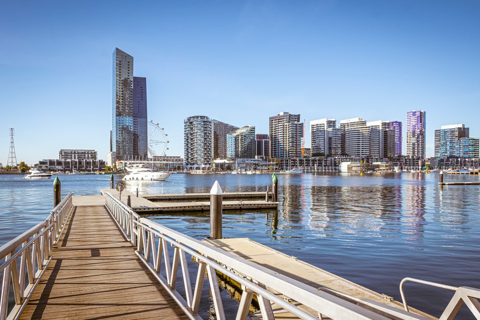 A panoramic view of Docklands featuring modern high-rise office buildings and residential flats made of glass and steel, reflecting sunlight and appearing clean and well-maintained. In the foreground, a marina with still water holding several white yachts and boats, their reflections clearly visible on the surface. The sky is bright blue with no clouds, and the scene is illuminated by natural daylight, emphasizing the sleek and tidy appearance of the waterfront area. The overall setting showcases a contemporary urban landscape with a focus on cleanliness and architectural detail, indicative of the professional cleaning services provided by Carpet Cleaning Docklands and aligned with the topic of avoiding common carpet cleaning mistakes in Docklands flats.