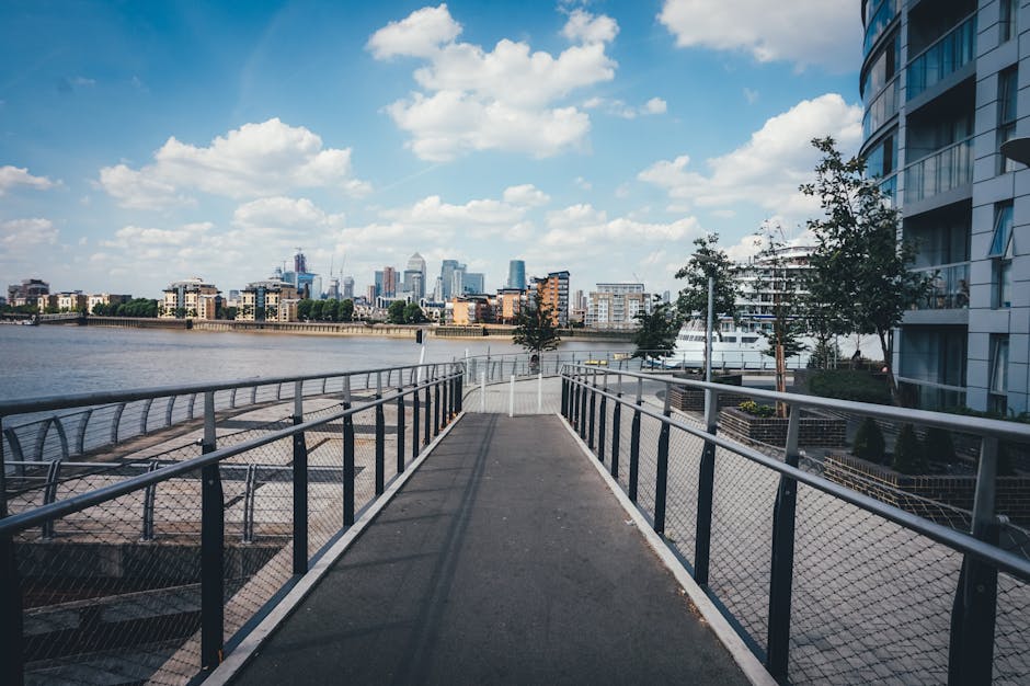 A paved pedestrian walkway with metal railing fencing along the River Thames in London, featuring modern residential buildings and trees on the right side, with a city skyline in the background under a partly cloudy blue sky, captured during daytime. The scene emphasizes urban outdoor surface infrastructure and landscaping, in a setting that highlights the area around Canary Wharf as referenced in the cleaning guide for flats and towers, with no visible cleaning equipment or surfaces. Carpet Cleaning Docklands specializes in surface cleaning and deep cleaning services for residential and commercial properties in the Docklands area.