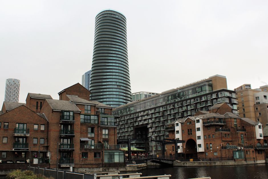 A daytime cityscape featuring a mix of residential buildings and modern high-rise towers in Canary Wharf, Docklands. The foreground displays red brick apartment complexes with multiple stories, glass balconies, and dark window frames, situated along a waterway with visible reflections on the surface. Behind them, a large, contemporary cylindrical skyscraper with horizontal glass bands dominates the skyline, alongside other modern buildings with glass facades and varied architectural styles. The scene is illuminated by natural daylight with overcast skies, highlighting the clean and well-maintained appearance of the surfaces and structures, aligning with the theme of professional surface cleaning and maintenance offered by Carpet Cleaning Docklands.