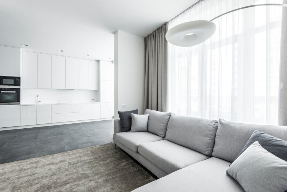 A bright and modern living room featuring a large light gray sofa with matching cushions, positioned on a neutral-colored area rug. Behind the sofa, there are floor-to-ceiling sheer white curtains allowing natural light to fill the space and side gray drapes. The room has a minimalist aesthetic with clean white walls and a contemporary ceiling light fixture. To the left, an open-plan kitchen with white cabinets, integrated appliances, and a gray tiled floor extends into the space, creating a seamless and hygienic environment. The surfaces appear clean and well-maintained, emphasizing a focus on surface cleaning, hygiene, and domestic maintenance, as promoted by Carpet Cleaning Docklands.
