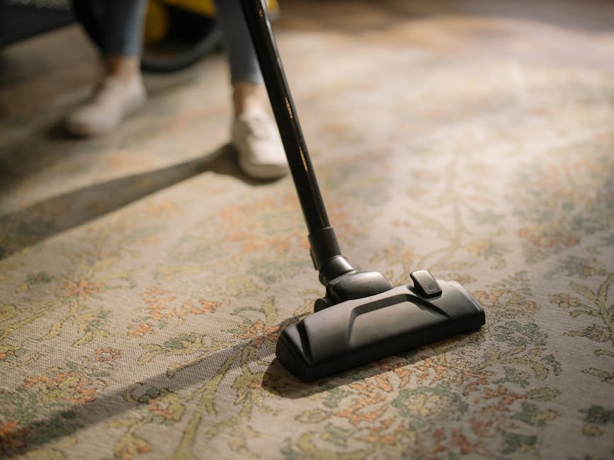 Close-up of a modern vacuum cleaner head actively cleaning a patterned area rug with floral motifs, situated in a well-lit room with natural light. The vacuum head is black with an adjustable height feature, and the wand extends upward out of view. The surface of the rug appears dust-free and well-maintained, indicating thorough surface cleaning. In the background, a person's legs and white sneakers are visible, suggesting an active domestic setting. This image typifies professional and domestic deep cleaning practices recommended by Carpet Cleaning Docklands, especially for period homes on the Isle of Dogs, emphasizing hygiene and the upkeep of delicate furnishings.