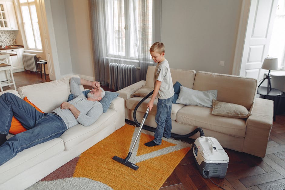 A young boy using a vacuum cleaner to deep clean a beige sofa in a bright living room with wooden flooring, white and light gray walls, and large windows with sheer curtains. An older man is lying on the sofa, relaxing with his eyes closed. The room features a small side table with a lamp, a radiator beneath the window, and decorative pillows on the sofa. The cleaning process is visibly focused on maintaining hygiene and surface cleanliness, exemplifying domestic cleaning practices in a period home on the Isle of Dogs, with attention to detailed surface and fabric sanitisation by Carpet Cleaning Docklands.