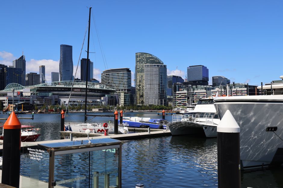 A panoramic view of Victoria Dock with modern high-rise buildings in the background under a clear blue sky, featuring a marina with various yachts and sailboats docked along the waterfront. The foreground shows a wooden dock with black and orange mooring bollards, and a glass railing with a sign indicating its location at Melbourne City Marina. The water reflects the boats and city skyline, emphasizing the clean and well-maintained environment typical of the area. The scene highlights the urban waterfront setting associated with the Royal Victoria Dock end of tenancy cleaning, underscoring the importance of surface cleaning and sanitisation in maintaining the pristine appearance of such commercial and residential spaces, as offered by Carpet Cleaning Docklands.