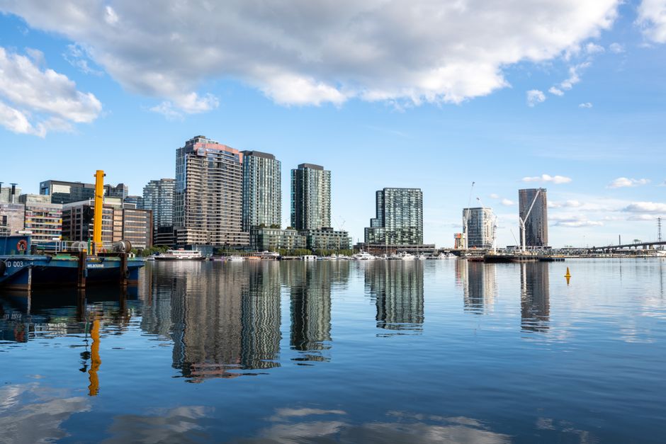 A waterfront view of the Royal Victoria Dock showcasing a modern skyline with multiple high-rise buildings made of glass and steel, reflecting on the calm water surface. The scene features a clear blue sky with scattered white clouds during daytime. In the foreground, part of a dock with a blue boat and yellow mooring post is visible, emphasizing the maritime environment. The photograph highlights the urban landscape cleaned and maintained for visual appeal, aligning with professional surface cleaning and cityscape hygiene standards, as promoted by Carpet Cleaning Docklands for end of tenancy cleaning and deep cleaning services.