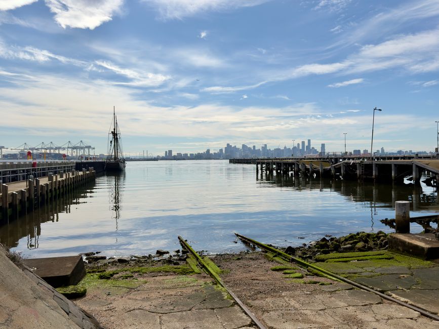 A panoramic view of Victoria Dock with modern high-rise buildings in the background under a clear blue sky, featuring a marina with various yachts and sailboats docked along the waterfront. The foreground shows a wooden dock with black and orange mooring bollards, and a glass railing with a sign indicating its location at Melbourne City Marina. The water reflects the boats and city skyline, emphasizing the clean and well-maintained environment typical of the area. The scene highlights the urban waterfront setting associated with the Royal Victoria Dock end of tenancy cleaning, underscoring the importance of surface cleaning and sanitisation in maintaining the pristine appearance of such commercial and residential spaces, as offered by Carpet Cleaning Docklands.