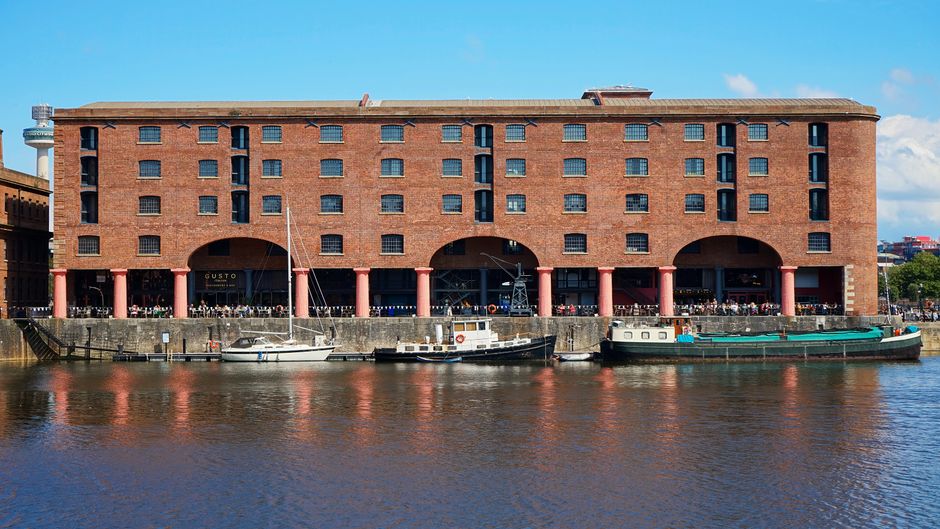 Exterior view of a large, rectangular brick building situated along a riverbank in Docklands, featuring multiple small rectangular windows on each floor and large arched openings at ground level supported by pink columns. Several boats are moored along the water's edge in front of the building, which has a flat roof and a modern industrial appearance. The scene is illuminated by bright sunlight, with a clear blue sky overhead and some greenery visible in the background. Carpet Cleaning Docklands specializes in surface cleaning and deep cleaning services, ensuring hygiene and maintenance for commercial and residential spaces in West India Quay and surrounding areas.