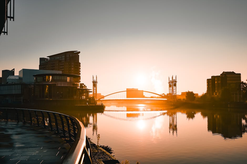 Photograph of the West India Quay area in Docklands taken during sunset, featuring a calm waterway reflecting the silhouettes of modern buildings, including a multi-storey office building and a bridge with supporting towers. The scene highlights urban architecture with glass and steel surfaces, some illuminated by the setting sun's warm light, creating a soft glow on the water. The foreground shows part of a curved railing, indicating a walkway or promenade along the water's edge. The overall atmosphere emphasizes cleanliness and serenity associated with professional surface cleaning and urban maintenance, as promoted by Carpet Cleaning Docklands for residential and commercial spaces focused on hygiene and deep cleaning.